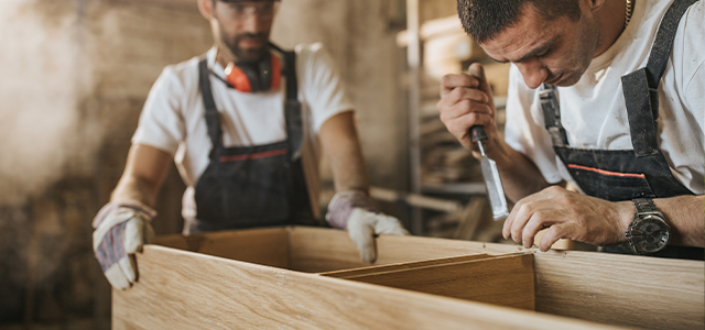 Zwei Schreiner arbeiten mit Werkzeugen an der Erstellung einer Kommode – Symbolbild für eine Gruppenunfallversicherung