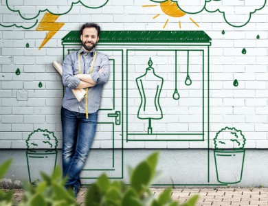 Mann mit einer Papierrolle und einem Maßband um den Hals gehängt steht vor seinem fiktiven an eine Wand gemalten Bekleidungsgeschäft