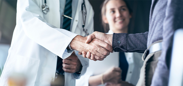 Händedruck zwischen Arzt und Patient, im Hintergrund eine Ärztin mit Klemmbrett – Symbolbild für eine Krankenversicherung