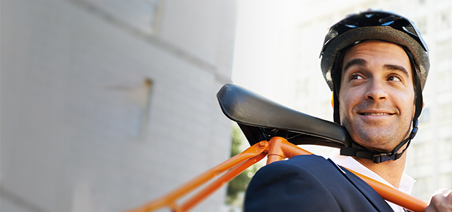 Mann im Anzug mit Fahrradhelm trägt sein Fahrrad mit der Stange über der Schulter – Symbolbild für eine Unfall invest Versicherung