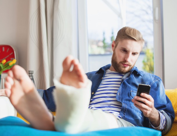 Mann liegt mit einem eingegipsten Bein auf der Couch und schaut auf sein Smartphone – Symbolbild für eine Unfallversicherung