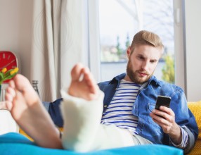 Mann liegt mit einem eingegipsten Bein auf der Couch und schaut auf sein Smartphone – Symbolbild für eine Unfallversicherung