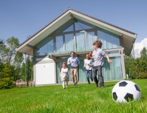 Familie spielt auf dem Rasen vor einem modernen Einfamilienhaus bei Sonnenschein – Symbolbild für eine Wohngebäudeversicherung