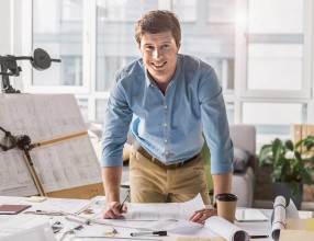 Ein jüngerer Mann, der als Selbstständiger arbeitet, steht an seinem Schreibtisch auf dem viele Papiere liegen