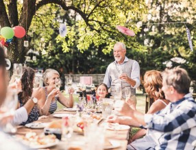 Ein älterer Mann hält bei einem Fest eine Rede an seine Familie, die am Tisch vor ihm sitzt und sie prosten sich mit Sekt zu – Symbolbild für Bestattungsvorsorge