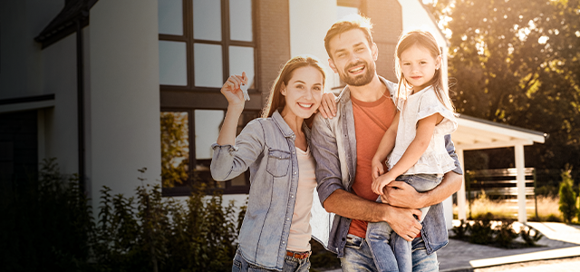 Eine Frau steht zusammen mit ihrem Mann, der die kleine Tochter auf dem Arm trägt, vor einem Haus und hält einen Schlüssel hoch – Symbolbild für Immobilienfinanzierung