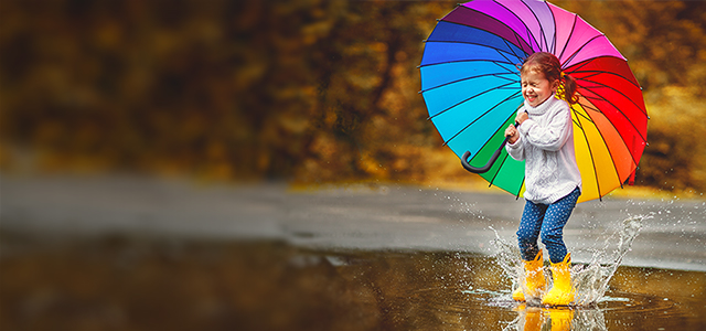 Kleines Mädchen spielt mit gelben Gummistiefeln und einem Regenschirm in den Farben des Regenbogens in einer Pfütze