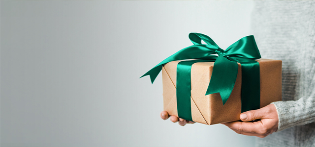 Ein Geschenkpaket mit einer grünen Schleife, wird von einer Person gehalten von der man nur die Hände sieht – Symbolbild für vermögenswirksame Leistungen