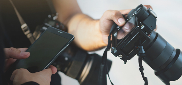 Fotograf hält eine Kamera, daneben prüft jemand ein Smartphone – Austausch digitaler Bilder – Symbolbild für die Kategorie Pressefotos